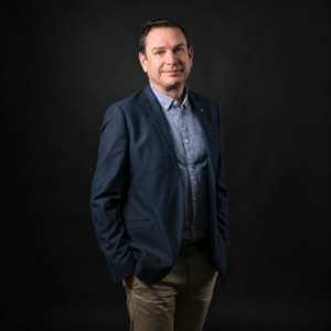 Julien HERBIN en veste bleu marine et chemise, souriant sur un fond sombre
